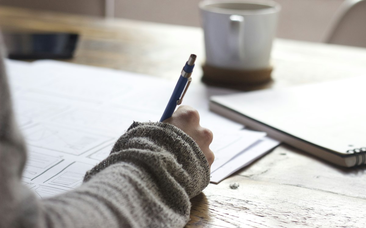 A person writing notes beside a cup of coffee.